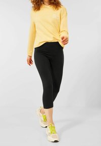 Yellow hooded sweatshirt with white text, paired with black capri leggings and light sneakers with lime green accents and mesh detailing.