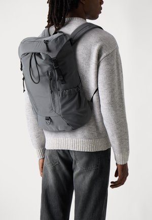 Grauer Nylon-Rucksack mit verstellbaren Trägern, einer vorderen Reißverschlusstasche, seitlichen Netztaschen und einem Tragegriff, getragen über einem grauen Strickpullover.