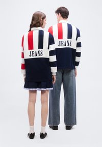 Δύο άτομα που κοιτούν προς τα πίσω φορούν ταιριαστά μακρυμάνικα πουκάμισα με κόκκινες, λευκές και ναυτικές ρίγες και την επιγραφή "JEANS" στην πλάτη.