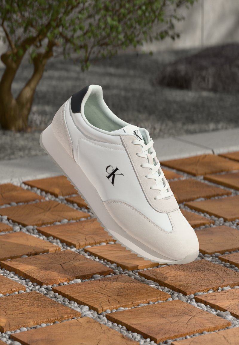 Witte en zwarte sneaker met veters en het "cK" logo die boven een houten tegel- en kiezeloppervlak zweeft.