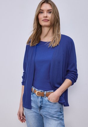 Jonge vrouw met blond haar, gekleed in een blauwe cardigan en top, lichtblauwe spijkerbroek en een beige riem, staand met één hand in haar zak.