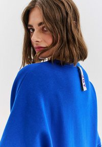 Modrá mikina z mäkkého materiálu, s okrúhlym výstrihom a výraznou nálepkou vzadu pri krku. Hladká textúra.