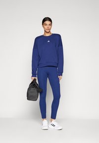Sweatshirt desportivo azul marinho com ombros caídos, combinado com leggings a condizer. Modelo segura uma mala de ginásio preta. Detalhes de ténis brancos visíveis.