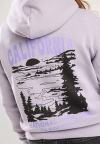 Levandulová mikina s černým grafickým designem západu slunce nad jezerem, se stromy a textem: "NÁRODNÍ PARK KALIFORNIE, USA." Text v purpurové barvě.