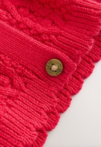 Rdeč pleteni kos oblačila z rebrasto teksturo, ki ima dekorativni vzorec z vrvico in medeninasto gumb z vgraviranim napisom.