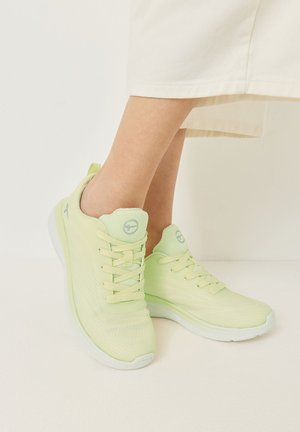 Lichtgroene mesh sneakers met veters gedragen door een persoon in crèmekleurige broek, staand tegen een effen lichte achtergrond.