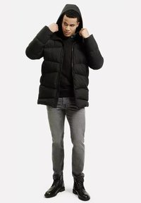 Czarna pikowana kurtka z kapturem, z uszytymi sekcjami. Nosi ją nad czarną bluzką, w zestawieniu z szarymi dżinsami i czarnymi sznurowanymi butami.
