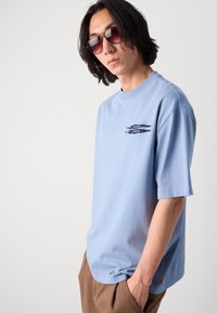 Tricou scurt, cu maneci scurte, de culoare albastru deschis, din bumbac, cu un croi lejer. Dispune de un design grafic de culoare albastru închis pe partea stângă a pieptului și o etichetă detaliată de culoare roșie.
