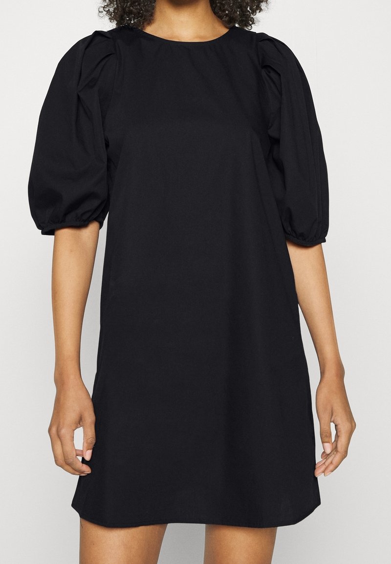 Femme portant une robe noire courte avec des manches bouffantes aux coudes et un col rond sur un fond clair uni.