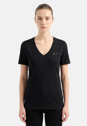 Giovane donna con pelle chiara e capelli raccolti dietro, che indossa una t-shirt nera con scollo a V e piccolo logo ricamato sul petto, in piedi su uno sfondo semplice.