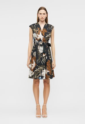 Frau steht und trägt ein schwarzes ärmelloses Kleid mit orange-, beige-farbenen Meereslebewesen-Mustern und gebundenem Taillengürtel, kombiniert mit weißen High-Heel-Sandalen.