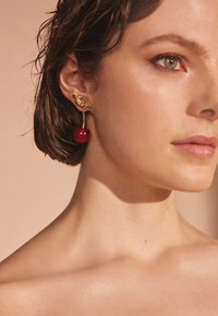 Náušnice v zlatej farbe s motívom čerešne, ktorá má lesklú červenú korálku, usadená na zakrivenej zlatej stonke. Pozadie je jemne ružové.