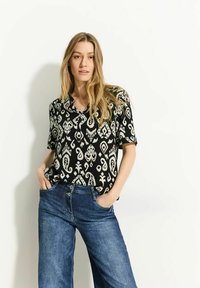 Sort kortærmet skjorte med hvide abstrakte mønstre, parret med højalmede blå jeans. Model poserer med hænderne i lommerne.