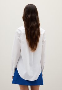 White button-up shirt with long sleeves and a curved hem, paired with a blue skirt. Smooth fabric, classic design, no visible patterns.