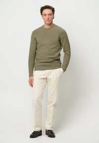 Maglione lavorato a maglia verde oliva con scollatura rotonda, abbinato a pantaloni color crema e mocassini scuri, che mettono in evidenza una silhouette aderente.