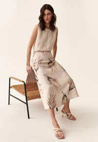 Beige lenvér ujjatlan top, térd hosszúságú szoknyával, amely barna levélmintákkal rendelkezik. A modellen bézs szandál van. Fonott szék háttér.