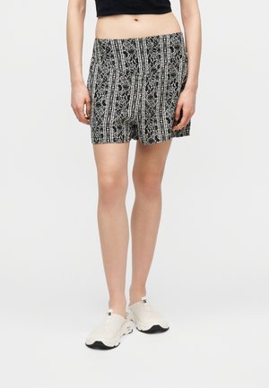 Γυναίκα που φοράει ασπρόμαυρα σορτς με ψηλή μέση και λευκά sneakers χωρίς κορδόνια, στέκεται μπροστά σε ένα μονόχρωμο φόντο.