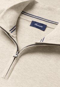 Beżowy sweter z dzianiny z półzamkiem i granatowymi paskami na kołnierzu, z naszywką w kolorze granatowym z białym napisem.