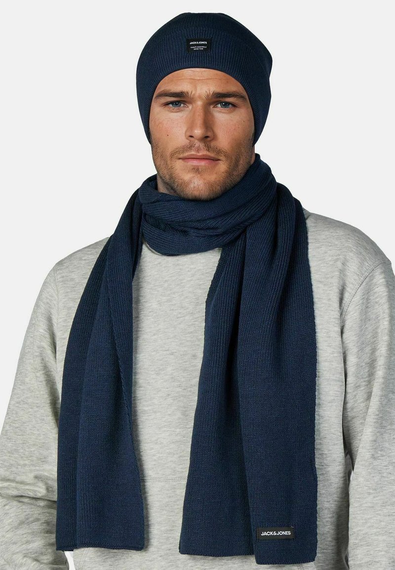 Marineblauwe gebreide muts en sjaalset, met geribbelde textuur en een logo-badge, gecombineerd met een grijze trui.