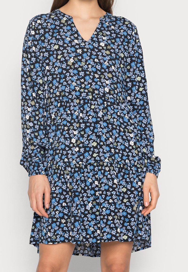 Femme portant une robe noire à manches longues avec un petit motif floral bleu, blanc et vert et un décolleté en V.