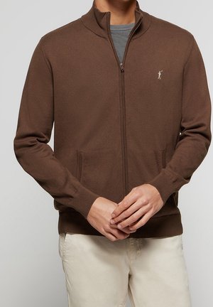 Homme portant un pull marron zippé sur une chemise grise avec un pantalon crème, mains jointes devant, sur un fond uni.