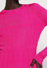 Top aderente a maniche lunghe in tessuto a coste di un brillante colore rosa. Presenta strisce verticali testurizzate e un collo rotondo. Le maniche arrivano fino al polso con polsini arricciati.
