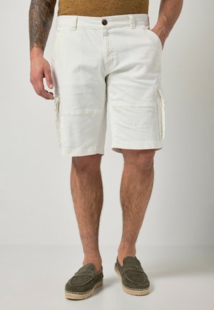 Mand iført hvide cargoshorts og olivengrønne slip-on sko står med den ene hånd i lommen.
