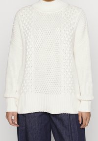 Pull blanc tricoté avec un col montant, des motifs texturés, des poignets et un ourlet côtelés. Le tissu semble doux avec des styles de tricot variés.