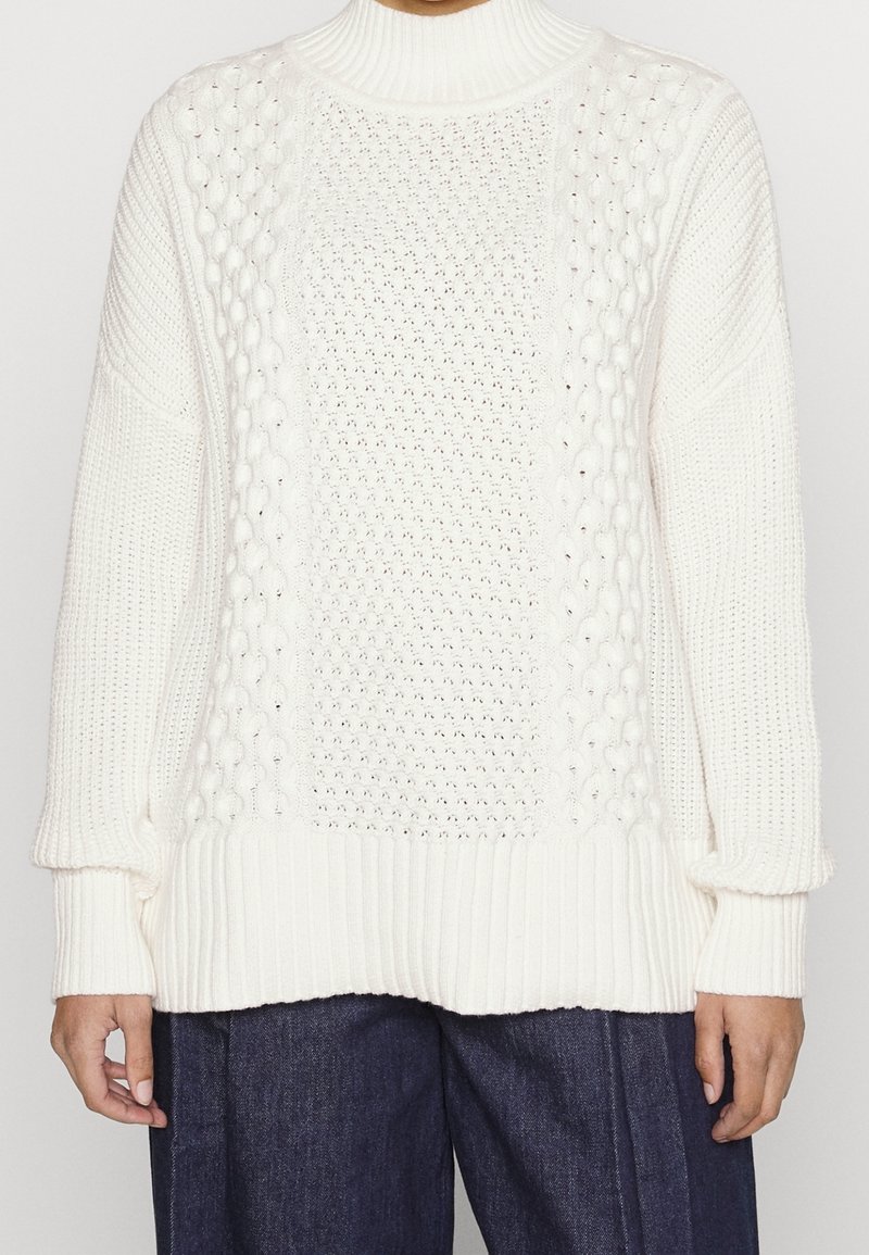 Pull blanc tricoté avec un col montant, des motifs texturés, des poignets et un ourlet côtelés. Le tissu semble doux avec des styles de tricot variés.