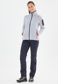 Graue Fleecejacke mit hohem Kragen, Reißverschluss-Taschen und roten Akzenten, kombiniert mit marineblauen wasserdichten Hosen und schwarzen Wanderschuhen.