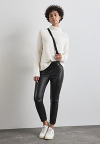 Överdimensionerad vit stickad tröja med hög krage, svarta byxor i konstläder och vita sneakers. Kort lockigt hår. Enkelt, avslappnat outfit.