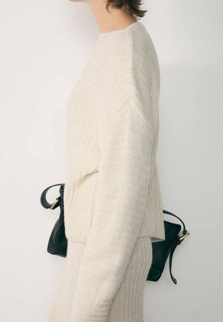 Femme portant un pull côtelé crème et une jupe, tenant un petit sac à main en cuir noir derrière son dos, sur un fond uni.