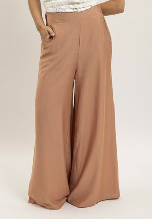 Femme portant un pantalon marron taille haute à jambes larges, les mains dans les poches, vue de la taille aux pieds sur un fond clair.