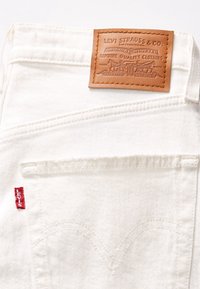 I jeans in denim bianco presentano un'etichetta in pelle marrone con testo in rilievo e un logo rosso Levi's sulla tasca posteriore; cuciture pulite esaltano il design.