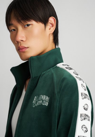 Giacca zip-up in velluto verde con colletto alto, presenta strisce laterali bianche stampate con il logo "Billionaire Boy Club", texture liscia.