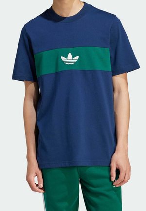 Mężczyzna w niebieskim t-shircie z zielonym poziomym paskiem i białym logo Adidas, w zestawieniu z zielonymi spodniami Adidas.