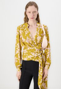 Langärmlige, senfgelbe Bluse mit pinkem Blumenmuster, die ein Wickeldesign und einen V-Ausschnitt aufweist. Kombiniert mit schwarzen hoch-taillierten Hosen.