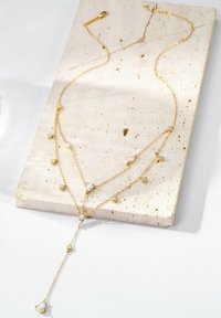 Gold-Schichtkette mit verschiedenen geschliffenen klaren Kristallen und einem Tropfanhänger. Ausgestellt auf einer strukturierten cremefarbenen Steinoberfläche.