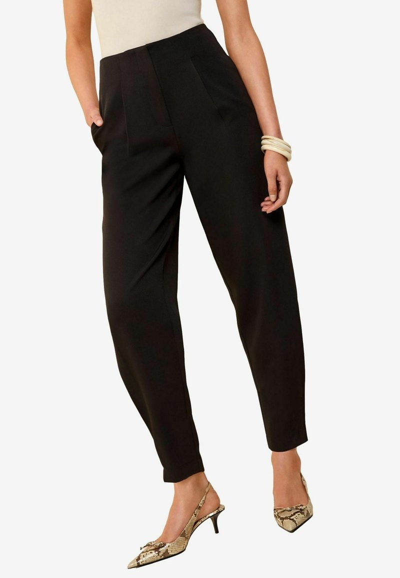 Femme portant un pantalon noir taille haute à jambes fuselées, un haut beige sans manches, des escarpins à lanières imprimé serpent, et des bracelets jonc crème au poignet droit.