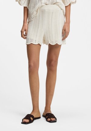 Person trägt weiße gestrickte Shorts mit wellenförmigen Kanten und schwarze Slide-Sandalen, steht vor einem schlichten hellen Hintergrund.