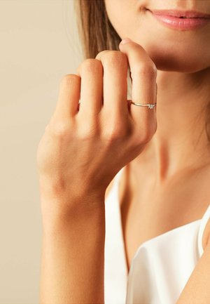 Goldring mit einem kleinen klaren Edelstein, getragen an einem schlanken Finger. Die Hautfarbe ist hell, mit natürlicher Textur und subtilen Details sichtbar.