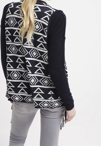 Svart och vit mönstrad väst med geometriska mönster, bärs över en långärmad svart topp, ihop med ljusgrå jeans som visar en fransdetalj.