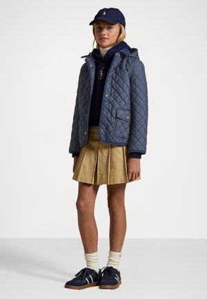 Giacca imbottita blu con cappuccio e tasche frontali, abbinata a una felpa blu, una gonna plissettata beige e sneakers blu con dettagli bianchi.
