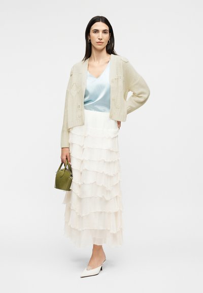 Γυναίκα με σκούρα μαλλιά που φοράει ένα κρεμ πλεκτό ζακέτα, ανοιχτό μπλε σατέν τοπ, στρωμένη λευκή φούστα, λευκές ψηλοτάκουνες και κρατά μια μικρή πράσινη τσάντα.