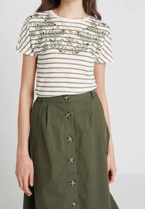 Femme portant un haut rayé blanc et olive à manches courtes avec broderie florale et une jupe olive à boutons sur le devant.