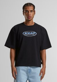 Black oversized t-shirt featuring a blue oval logo with the text "KANI" on the chest. Fabric appears soft, with a relaxed fit and short sleeves.