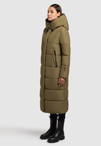 Olivgrön puffjacka med hög krage, dragkedja sidoficka och quiltat mönster. Bärs med svarta ankelstövlar och åtsittande byxor.