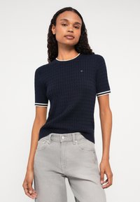 Ναυτικό μπλε κοντομάνικο πλεκτό πουκάμισο με υφασμάτινο σχέδιο και λευκή ριγέ κολάρο και μανίκια. Μικρό λογότυπο στο στήθος, συνδυασμένο με ανοιχτό γκρι παντελόνι.