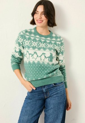 Frau trägt einen gestreiften Pullover in Türkis und Weiß mit Pinguinen und geometrischen Formen, kombiniert mit blauen Jeans, lächelt und schaut zur Seite.