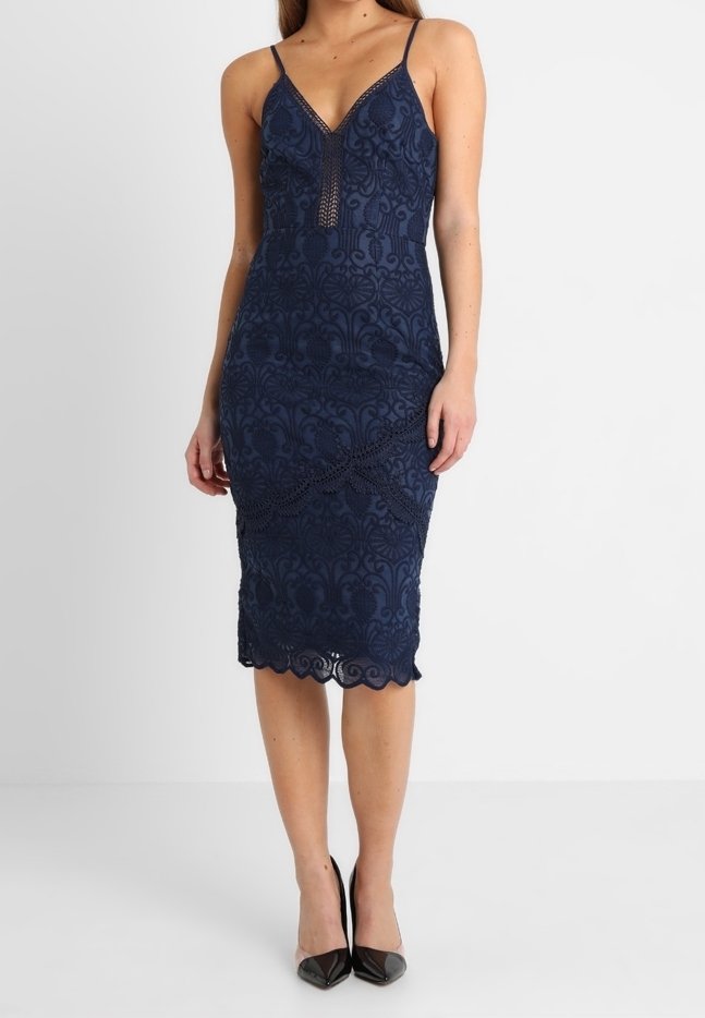 Femme portant une robe midi en dentelle bleu marine ajustée avec des bretelles spaghetti et des escarpins noirs à bout pointu, debout sur un fond blanc.